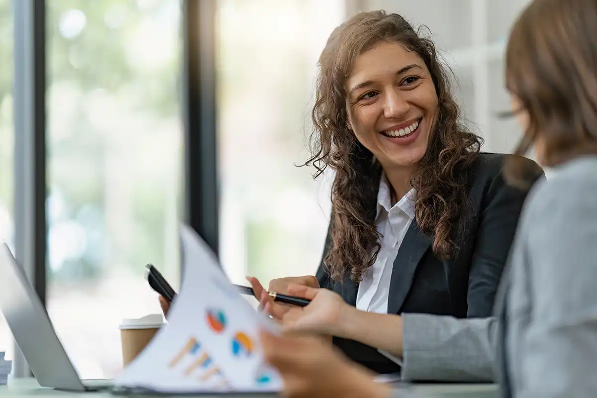 Two businesswomen discussing over documents and a laptop in a brightly lit office, one woman is smiling at her colleague about association management financials.