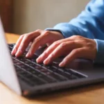 Hands typing on a laptop keyboard, researching HOA management services, while placed on a wooden table.